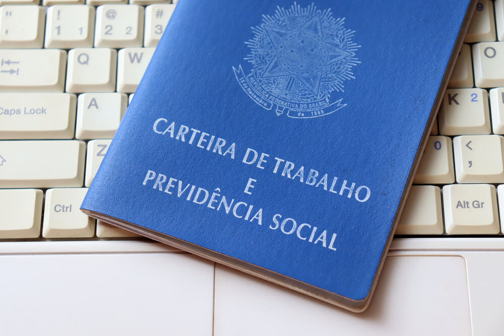 Imagem de uma Carteira de Trabalho e Previdência Social (CTPS) em cima do teclado de um computador branco.