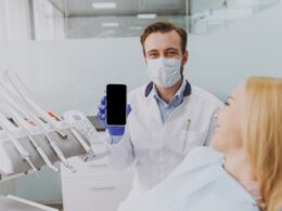 Dentista mostrando smartphone com paciente na cadeira odontológica.