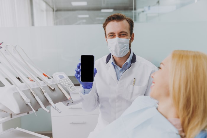 Dentista mostrando smartphone com paciente na cadeira odontológica.